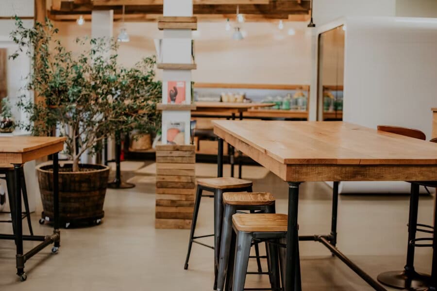 Tables en bois industriel et tabourets en métal dans un espace de type café décoré de plantes et d’un éclairage chaleureux.