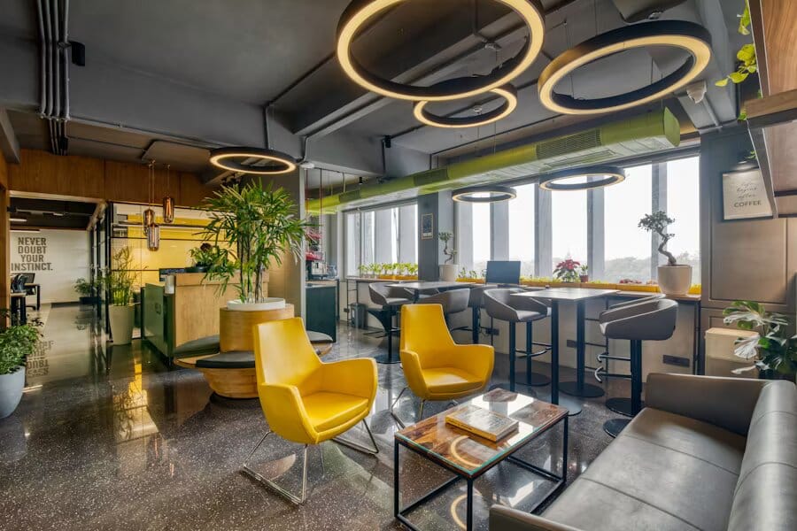 Salle de détente de bureau avec fauteuils jaunes, luminaires circulaires suspendus et plantes vertes près des fenêtres